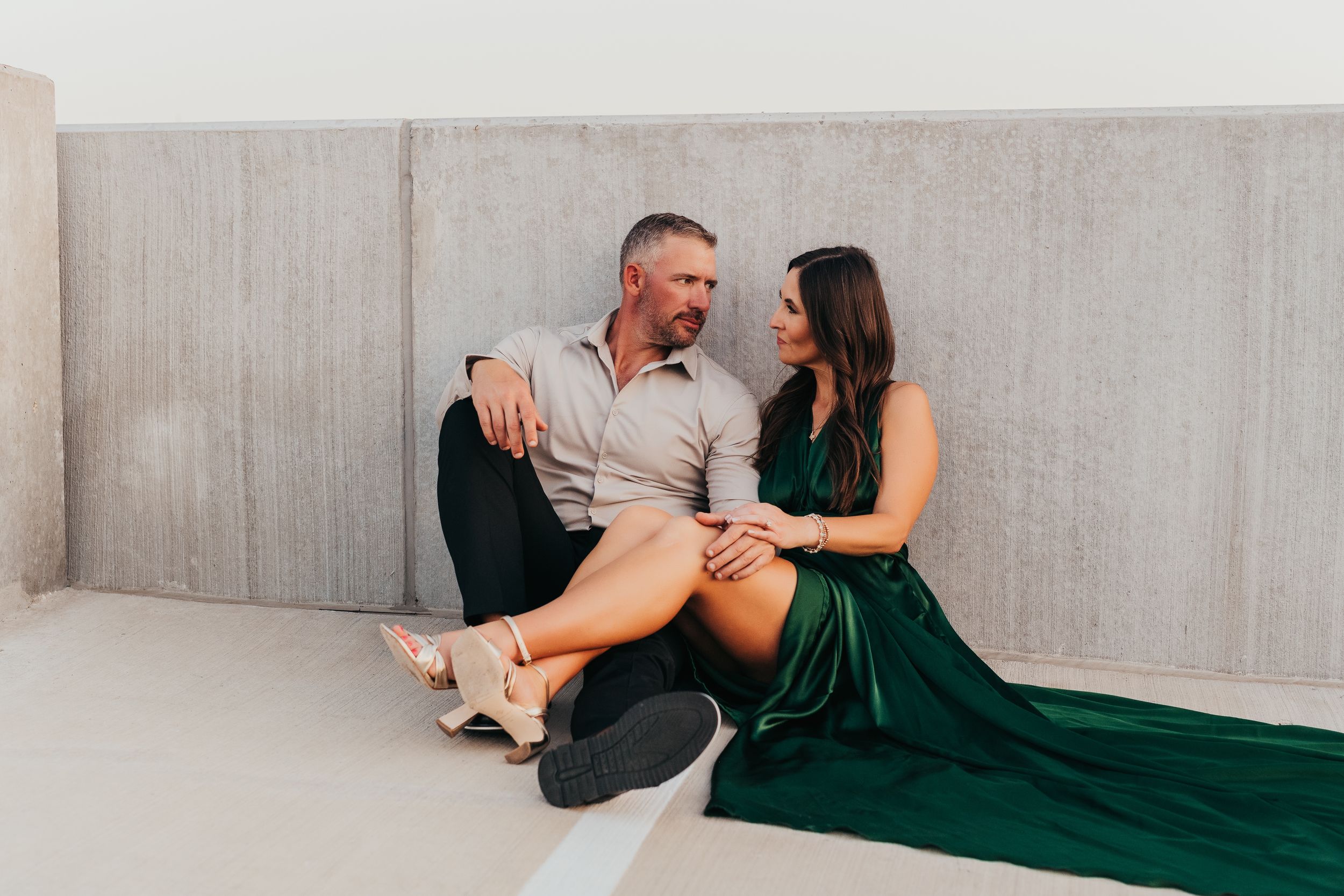 Couple in elegant attire sitting together against concrete wall, woman in green dress, man in beige shirt.