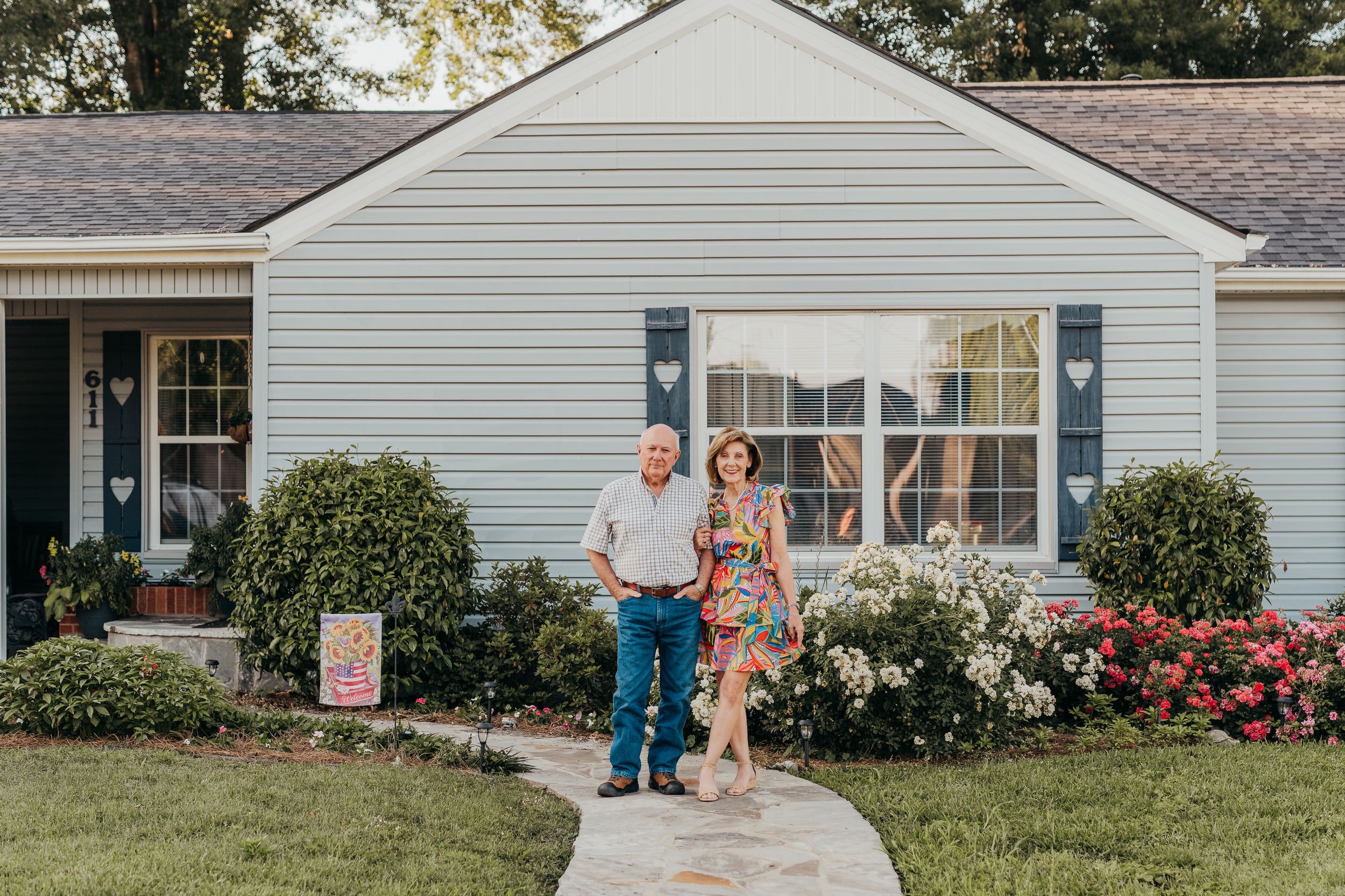 Couple standing with dog on walkway in front of charming blue ranch home with colorful flower gardens.