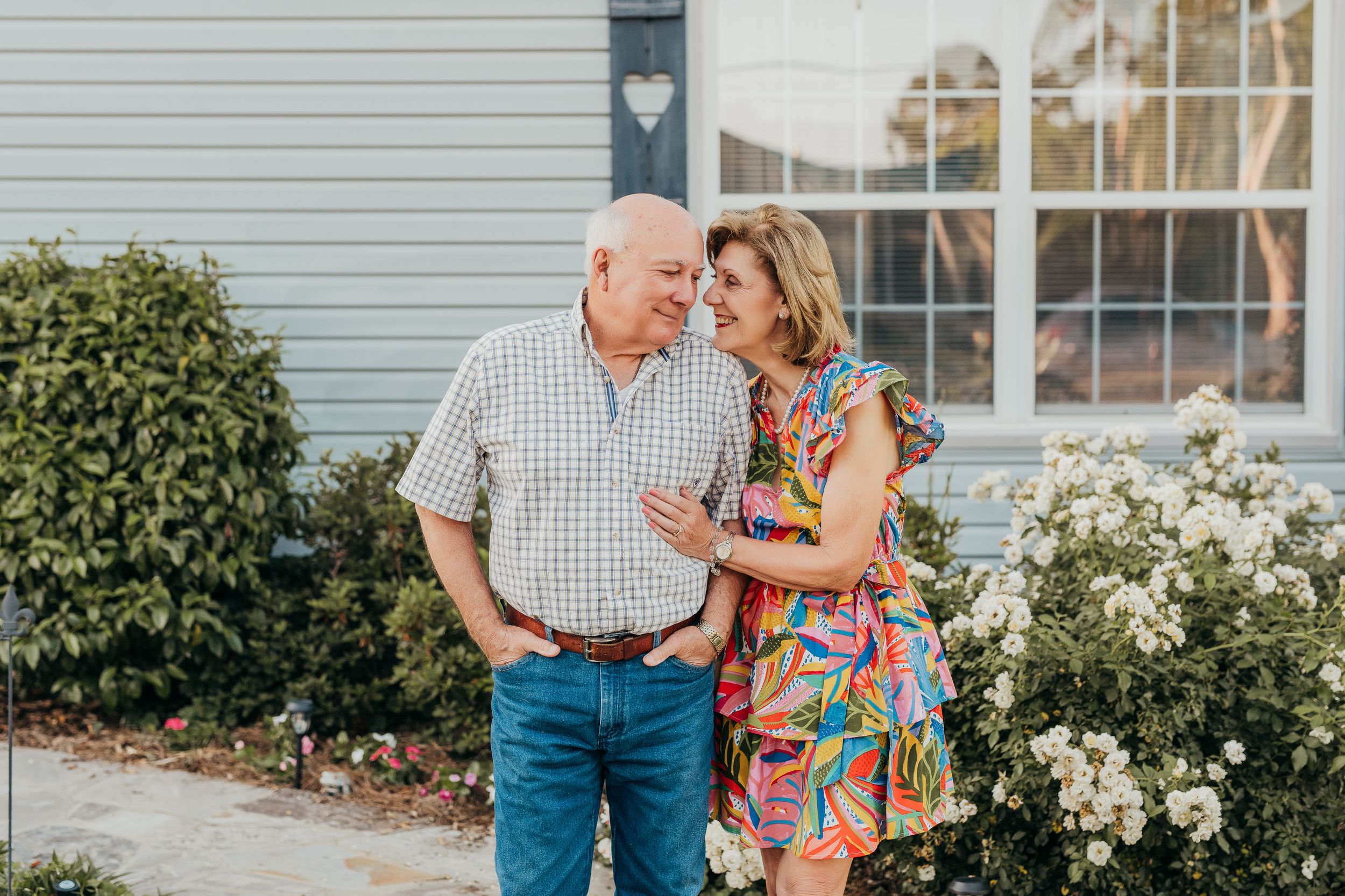 Older couple smiling at each other outside a blue house surrounded by white flowers and greenery.