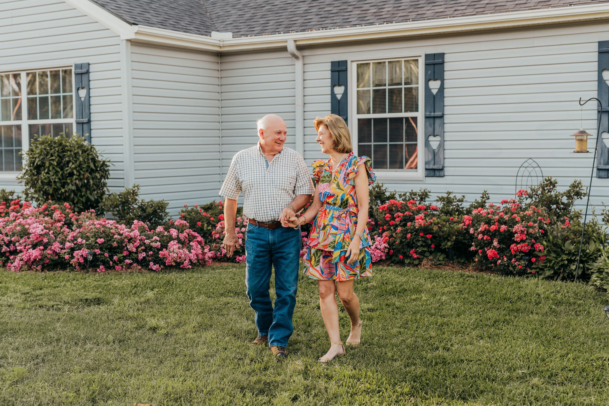 Elderly couple walking hand in hand on green lawn beside colorful pink rose bushes in front of white house.