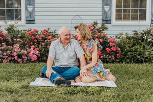 Happy elderly couple sitting on a blanket in the grass in front of a house with pink roses.