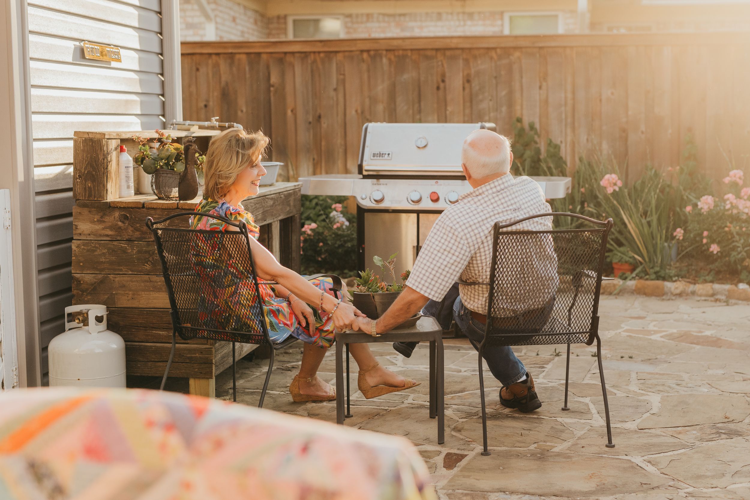 Elderly couple holding hands and relaxing together on a patio with a grill in the background.