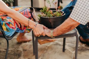 Close-up of an elderly couple holding hands on a stool beside a potted succulent plant.