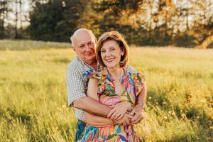 Smiling elderly couple embracing in a sunlit meadow with golden trees in the background.