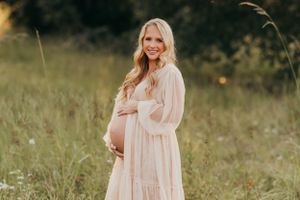 Expectant mother in cream dress stands in tall grass during golden hour.