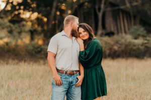 Romantic couple embracing in a field during golden hour, she in green dress and he in casual attire.