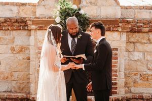 An outdoor wedding ceremony scene against a rustic stone and brick wall with floral decorations.