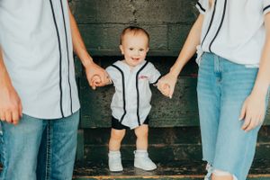 Small toddler in white baseball outfit holding hands with parents while standing on wooden steps.