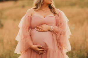 Expecting mother in flowing pink tulle dress poses in golden field during sunset maternity photoshoot.