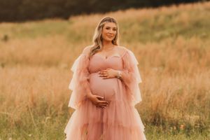 Maternity dress inspo- blush pink tulle gown stands in golden wheat field during sunset.