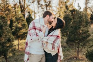 Couple wrapped in plaid blanket shares intimate moment among evergreen trees at golden hour.