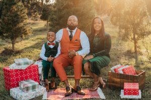 Family in festive attire poses on blanket with wrapped Christmas presents in wooded setting.