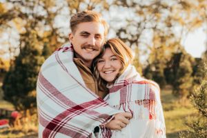 Couple wrapped in red and white plaid blanket sharing warm embrace in autumn setting.