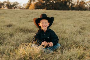 Child in black cowboy hat and denim sitting in tall grass during golden hour.