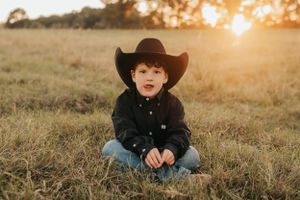 Young cowboy sitting cross-legged in a field at sunset wearing black hat and button-down shirt.