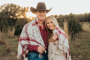 Couple in western attire and plaid blanket poses together in rustic outdoor setting.