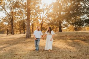 Couple holding hands while walking through a sunlit wooded area in casual attire.