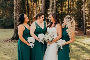 Bridesmaids in emerald green dresses hold white bouquets while posing in a forest setting.