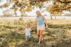 Adult and toddler walking together through sunlit field with autumn trees.