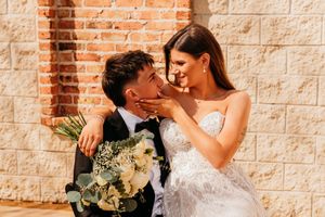 A romantic wedding couple sharing an intimate moment against a rustic brick wall with a white floral bouquet.
