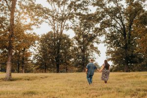 Two people walking through a golden meadow with tall trees in the background at sunset.