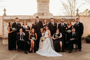 Wedding party in formal black attire poses together on stone terrace with vintage architectural backdrop.