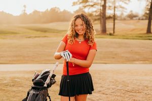 Golfer in red shirt and black skirt smiles while retrieving club from golf bag during golden sunset hour.