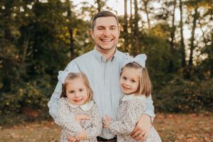 A dad and his daughters pose together outdoors during an autumn photo session.