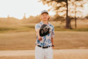 A person holds up a golf disc at golden hour on a grassy field with sunlit trees in the background.