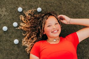 A person in a red shirt lying on grass with golf balls playfully scattered around them.