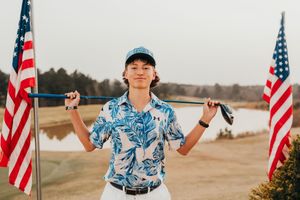 Golfer in Hawaiian print shirt stands between American flags on sandy terrain near water.