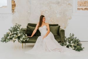 An elegant bride in a lace wedding gown sits on a dark green velvet couch adorned with lush greenery arrangements.