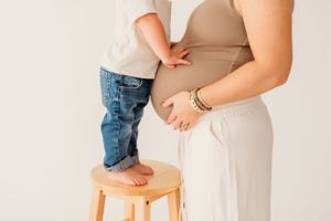 Child in jeans stands on wooden stool next to pregnant woman wearing beige top and white pants.