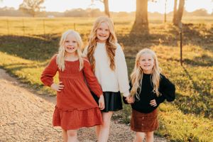 Three sisters in autumn outfits pose on a dirt path at sunset.
