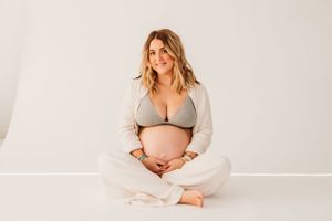 Pregnant woman in grey sweater and white pants sits cross-legged on white floor.