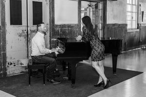 A black and white photograph shows two people near a grand piano in an old warehouse-like space.
