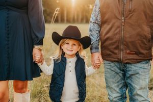 Son wearing a cowboy hat holds hands with parents in a sunlit field.