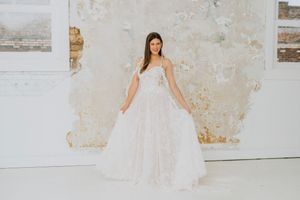 A bride stands in an elegant white ballgown against a minimalist distressed wall backdrop in a bright studio space.