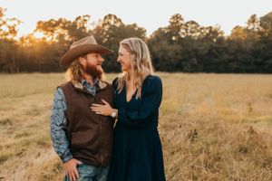 Couple embracing in a field at sunset with person wearing brown vest and cowboy hat.