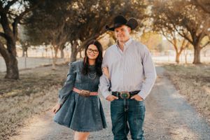 Engaged couple in denim and western wear pose together on a tree-lined path at sunset.