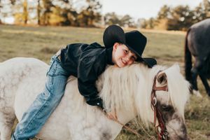 Child in western attire laying on a white horse in a grassy field at sunset.