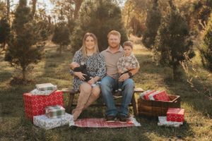 Family sits on picnic blanket surrounded by wrapped Christmas gifts at sunset in pine forest.