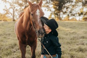 Young cowboy in black hat and shirt gently petting a brown horse in a pastoral setting.