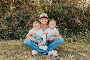 A person in a baseball cap and jeans sits cross-legged on grass with two young toddlers in matching blue outfits.