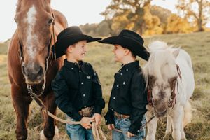 Two children in cowboy hats stand between brown and white horses in a field.