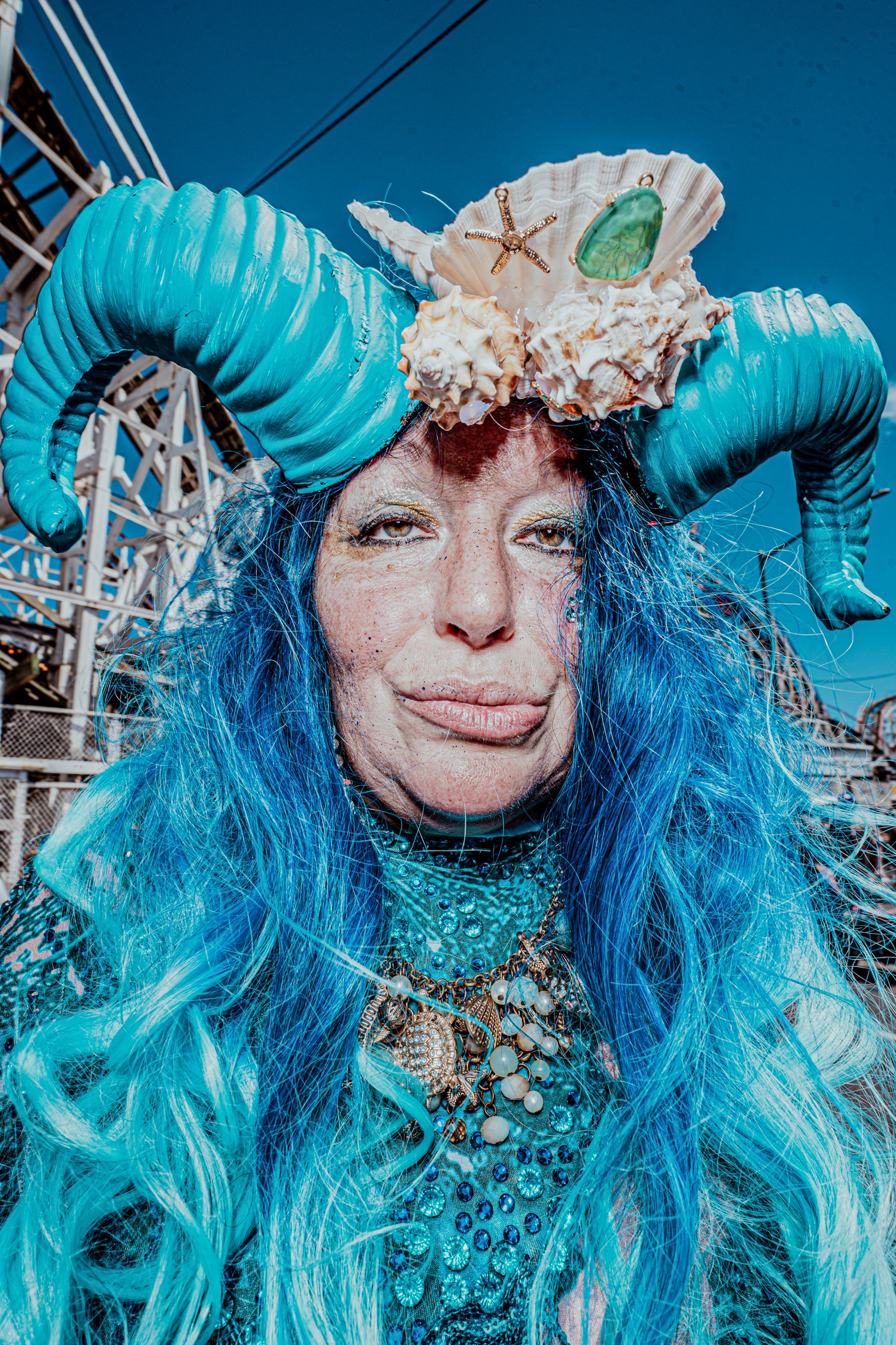 Woman in elaborate teal mermaid costume with blue hair and shell horns at Coney Island parade.