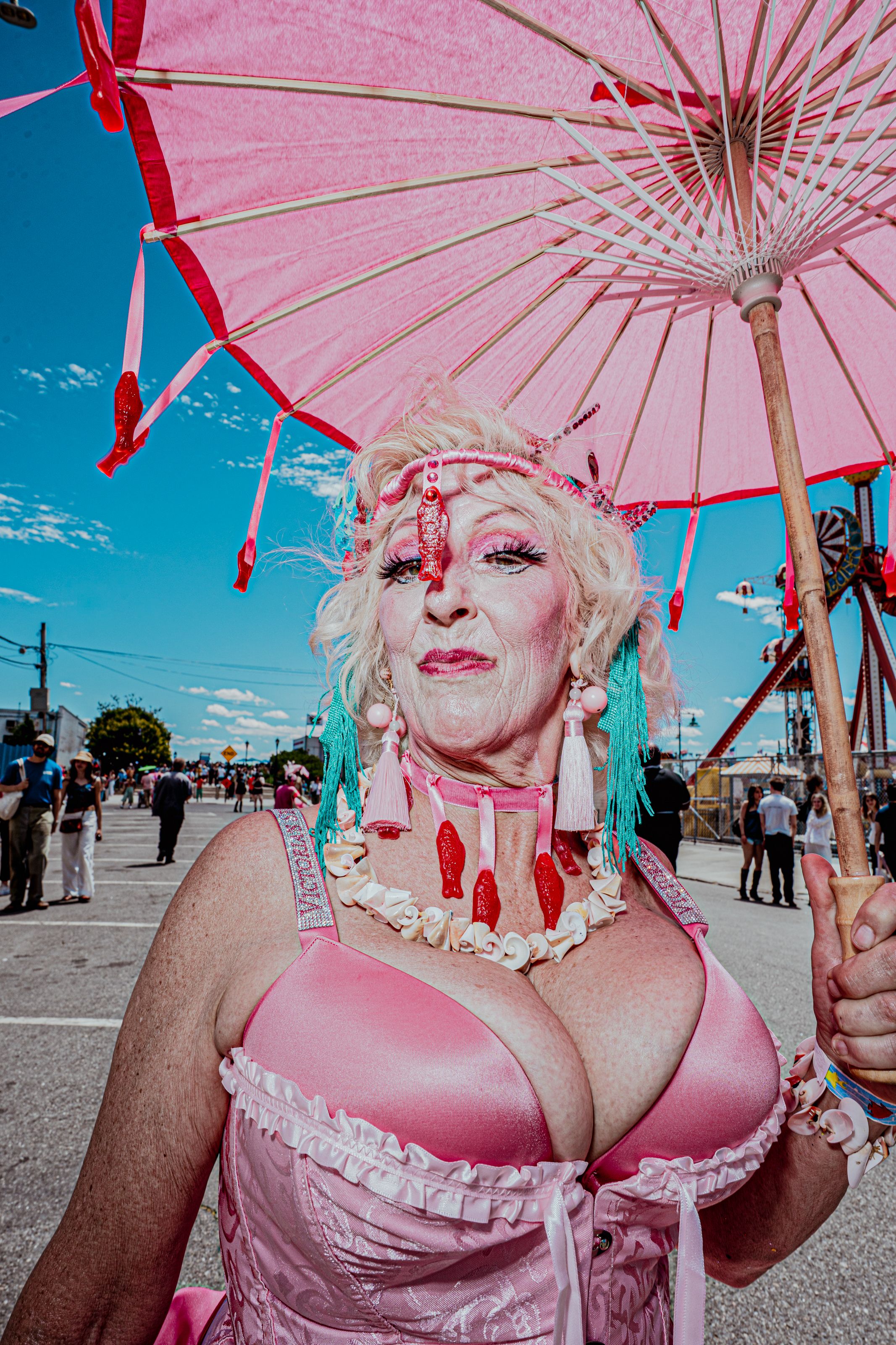 Senior woman in pink costume with large décolletage holds pink parasol at outdoor festival.