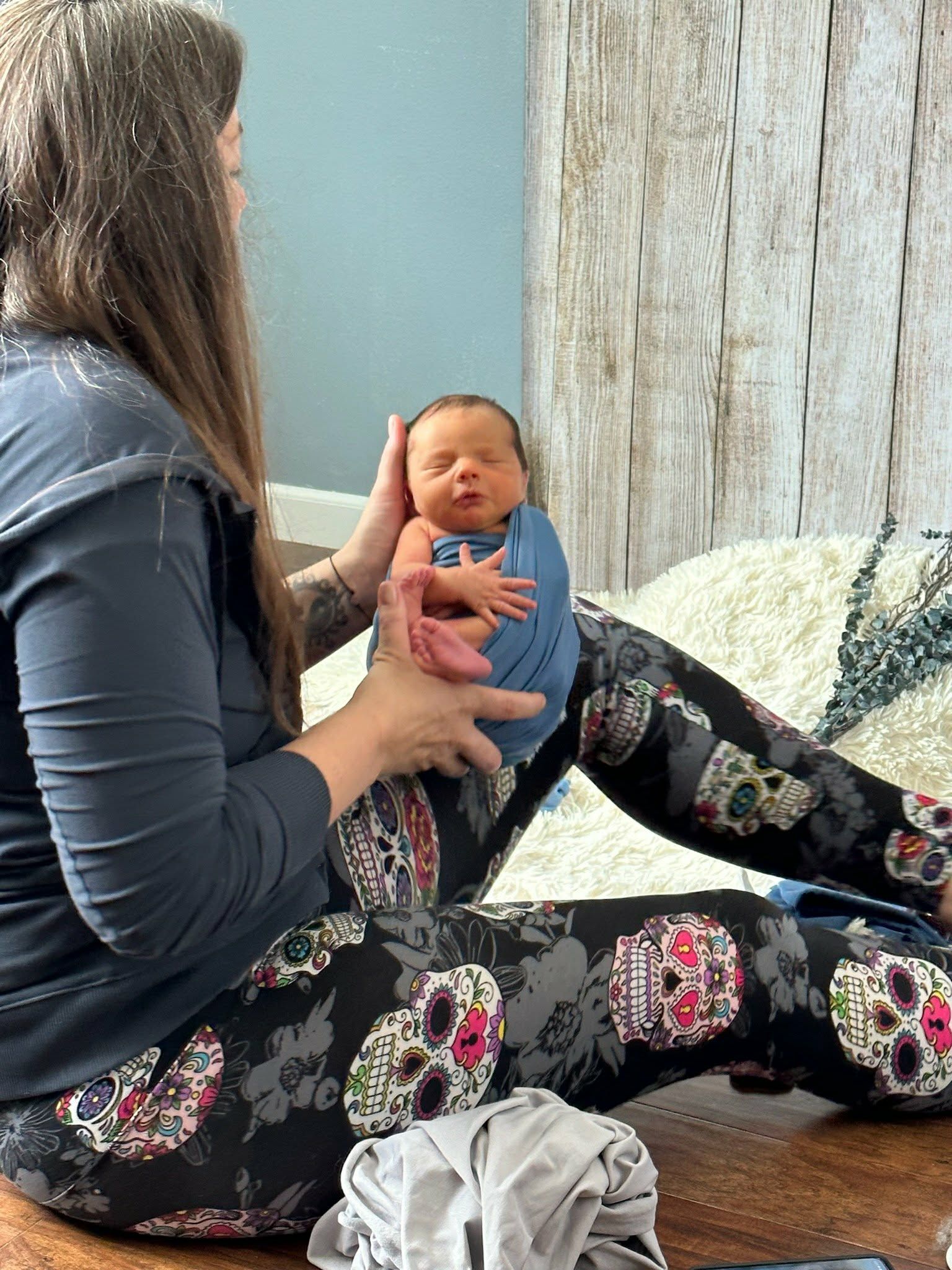 A woman in skull-print leggings gently holds a swaddled newborn baby while sitting on the floor.