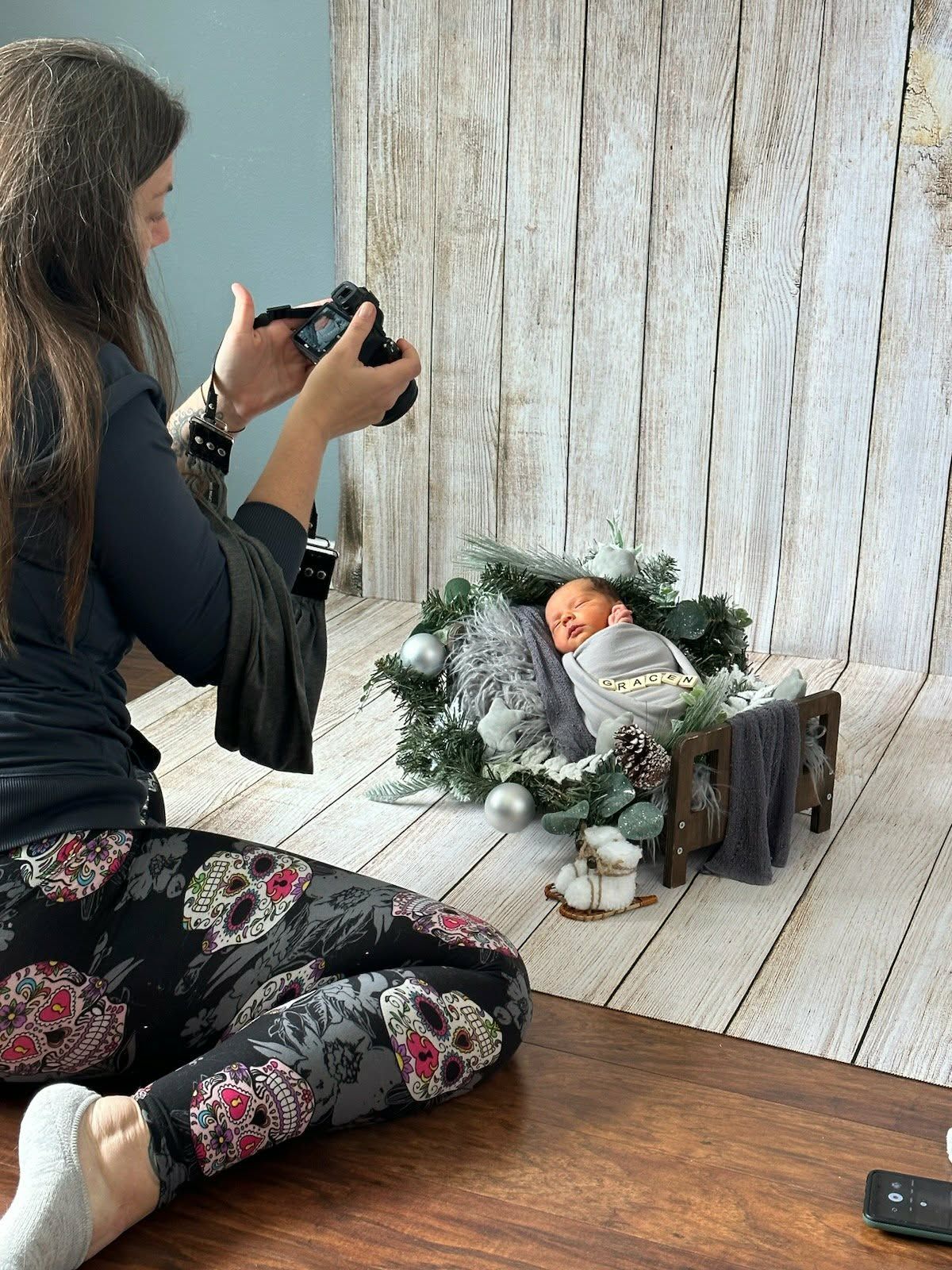 Photographer sitting cross-legged capturing newborn baby in festive Christmas wreath prop during professional studio photo session.