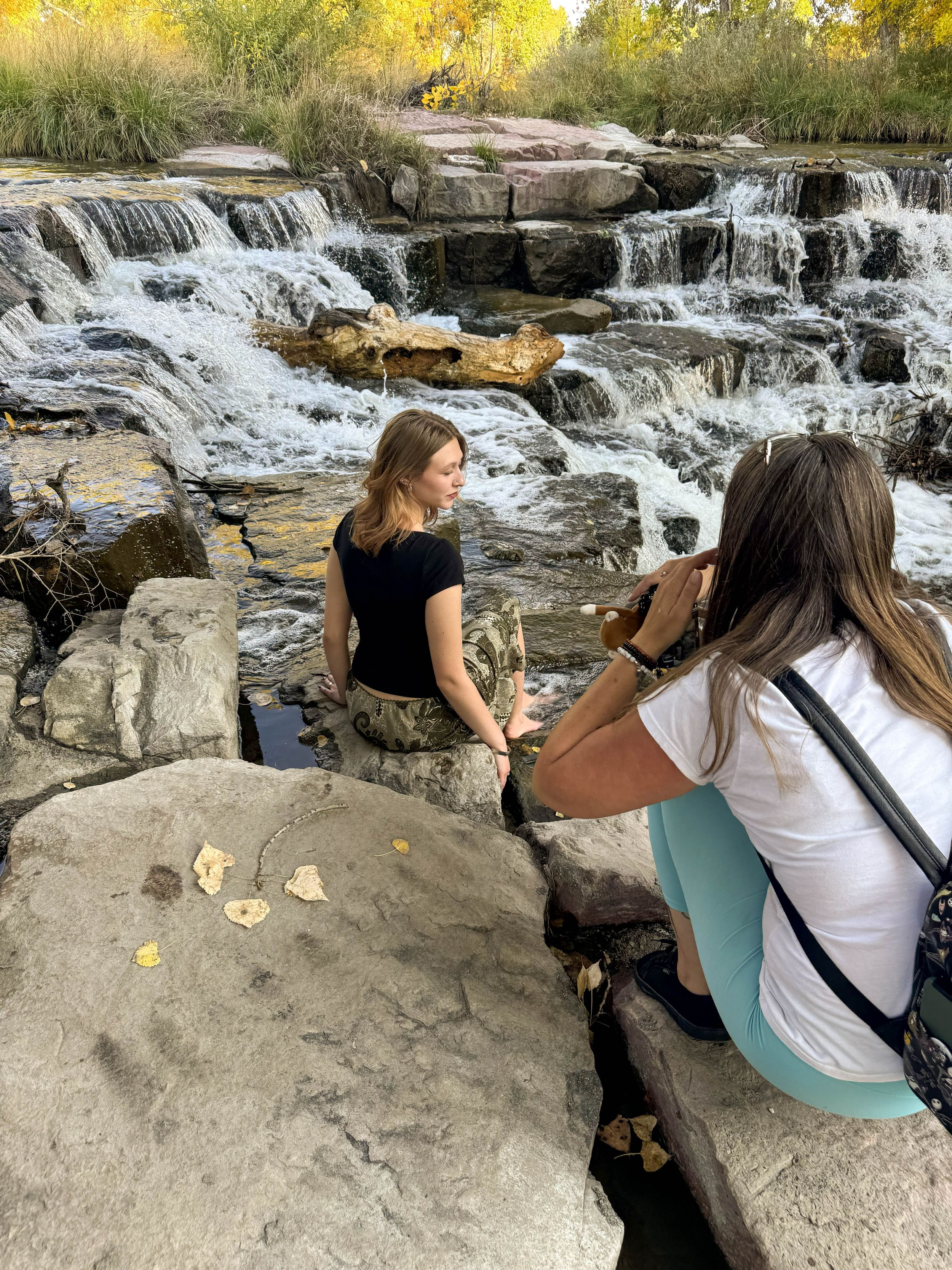 Woman being photographed by a friend near a cascading waterfall surrounded by autumn foliage and rocky terrain.